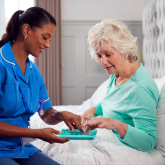 A nurse in blue uniform assists an elderly woman in a green shirt, sitting on a bed in a well-lit room.