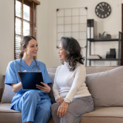 Healthcare professional in blue scrubs discussing medical information with a patient on a couch in a cozy living room.