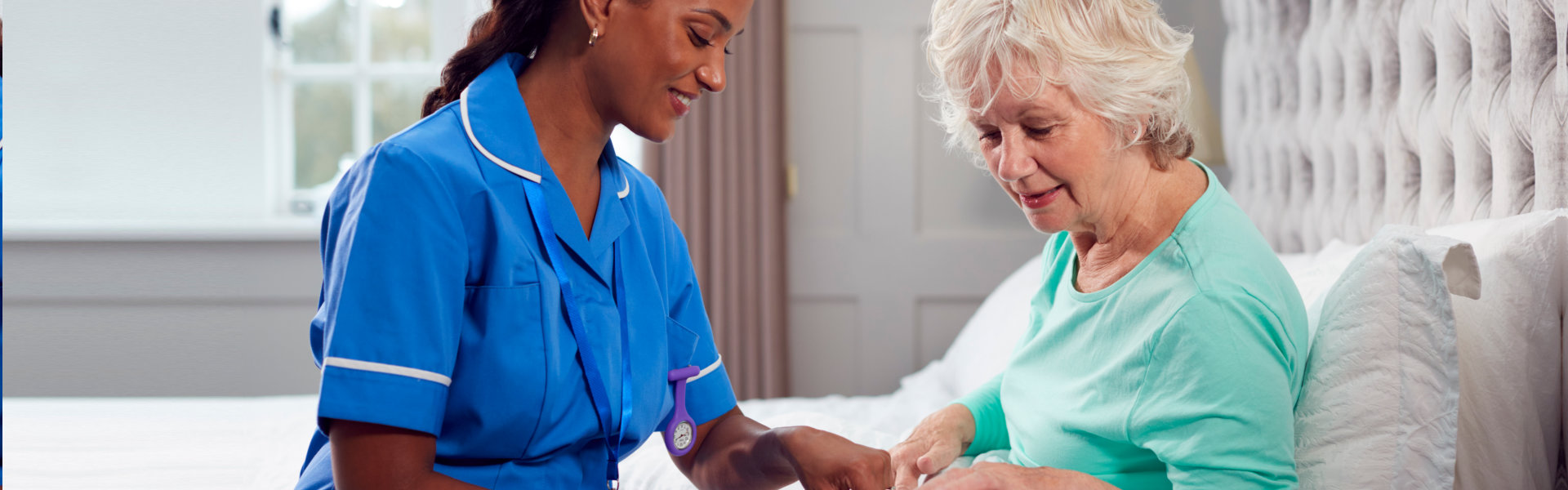 A nurse in blue uniform assists an elderly woman in a green shirt, sitting on a bed in a well-lit room.