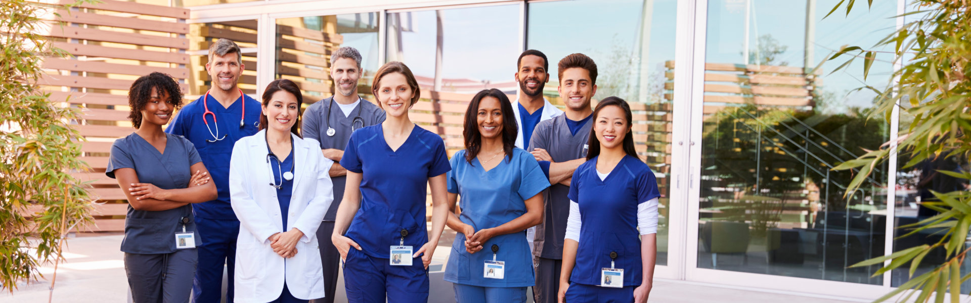 Diverse group of healthcare professionals in scrubs and lab coats standing together outside a medical facility.